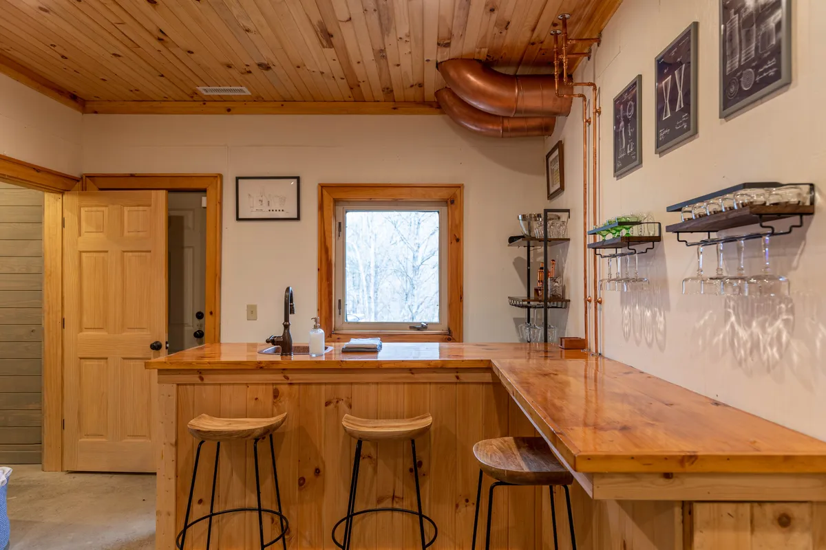 A cozy bar nook featuring a wooden bar counter with high stools, a window looking out onto the trees, and decorative wall art. The setting is perfect for sipping a crafted cocktail or a morning coffee while enjoying the view.