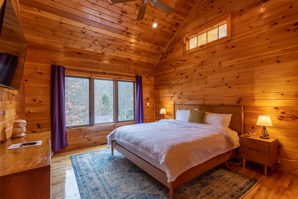 This image captures a serene bedroom with a plush bed, large window framing tranquil nature views, a decorative rug, a nightstand, and a wall-mounted TV. The soft light filtering through the window accentuates the calming atmosphere, perfect for relaxation or reading.