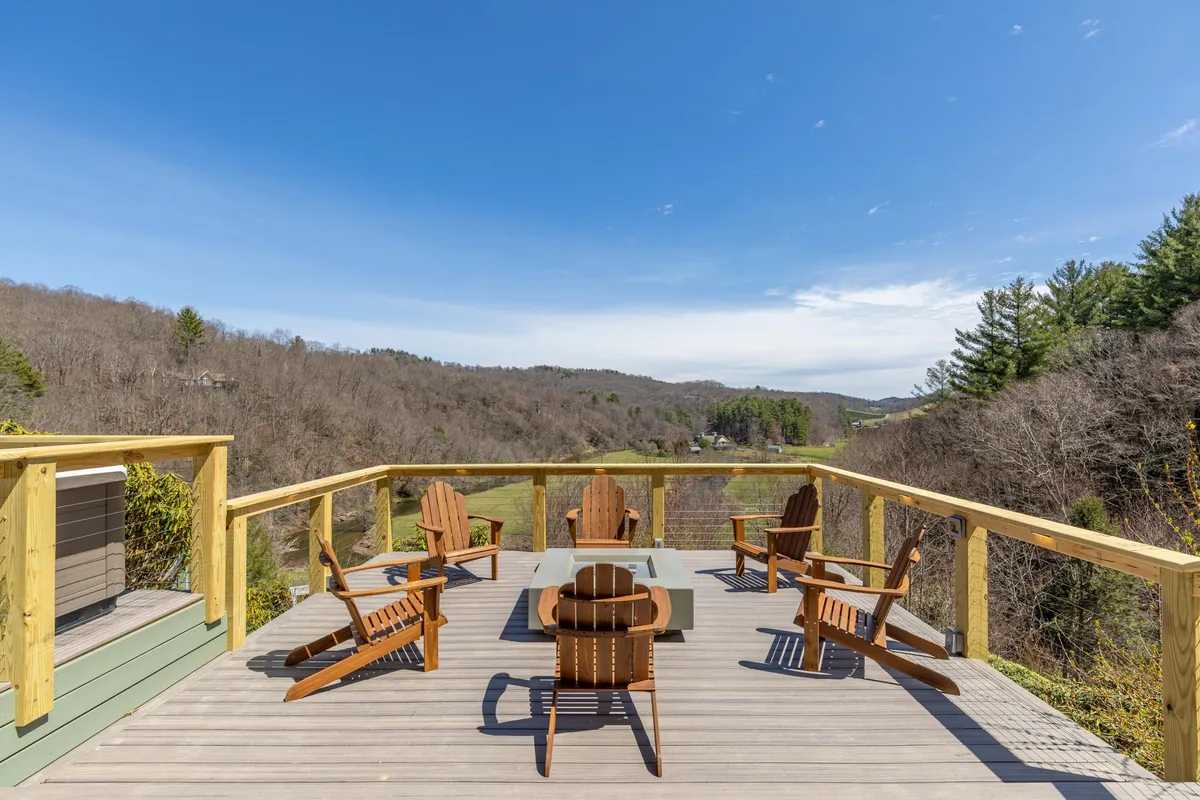 A spacious wooden deck is furnished with Adirondack chairs and a table, overlooking a sweeping mountain view. The setup invites relaxation, stargazing, and outdoor dining, with the expansive landscape changing hues throughout the seasons.
