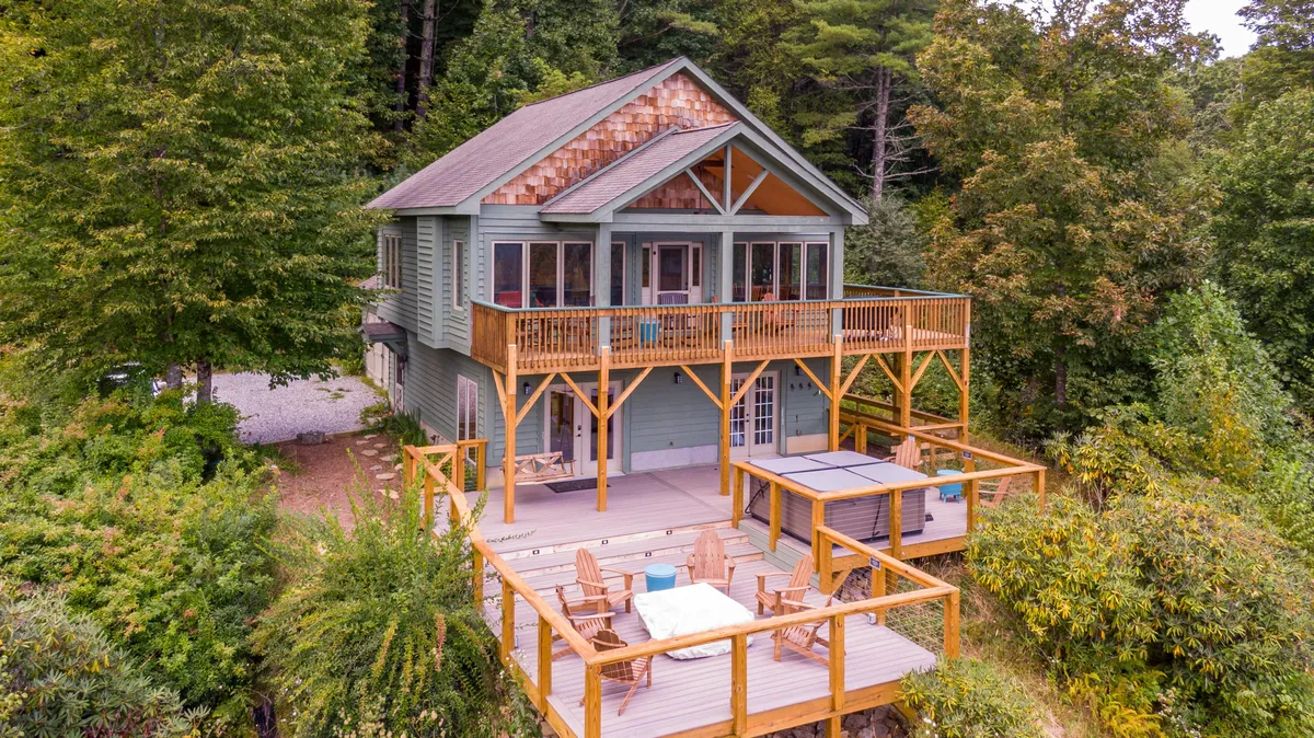 The image shows the cabin's spacious wooden deck nestled amid lush greenery, with a bubbling hot tub, large windows reflecting the surrounding trees, and panoramic views of the Appalachian countryside. The tranquil river can be glimpsed below, and the scene is bathed in the warm glow of sunset, inviting relaxation and outdoor enjoyment.