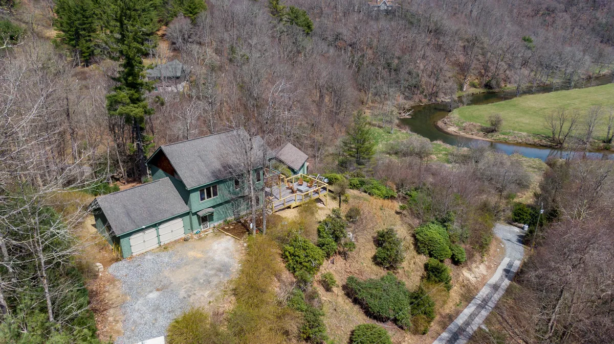 An exterior view of the house with a spacious deck overlooking the nearby river, surrounded by vibrant greenery. The deck is furnished for relaxation or outdoor dining and offers immersive views of the river and hills beyond, creating a serene and private retreat.