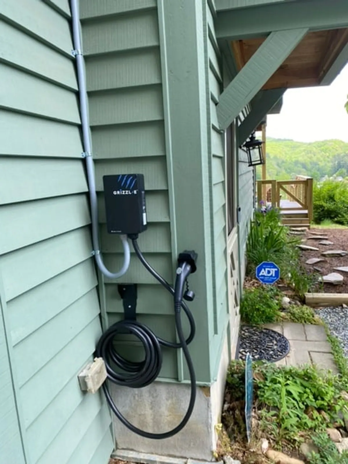 The home's EV charging station is positioned beside green siding and lush landscaping, highlighting eco-friendly amenities. The area is neat and easily accessible, blending modern convenience with natural surroundings.