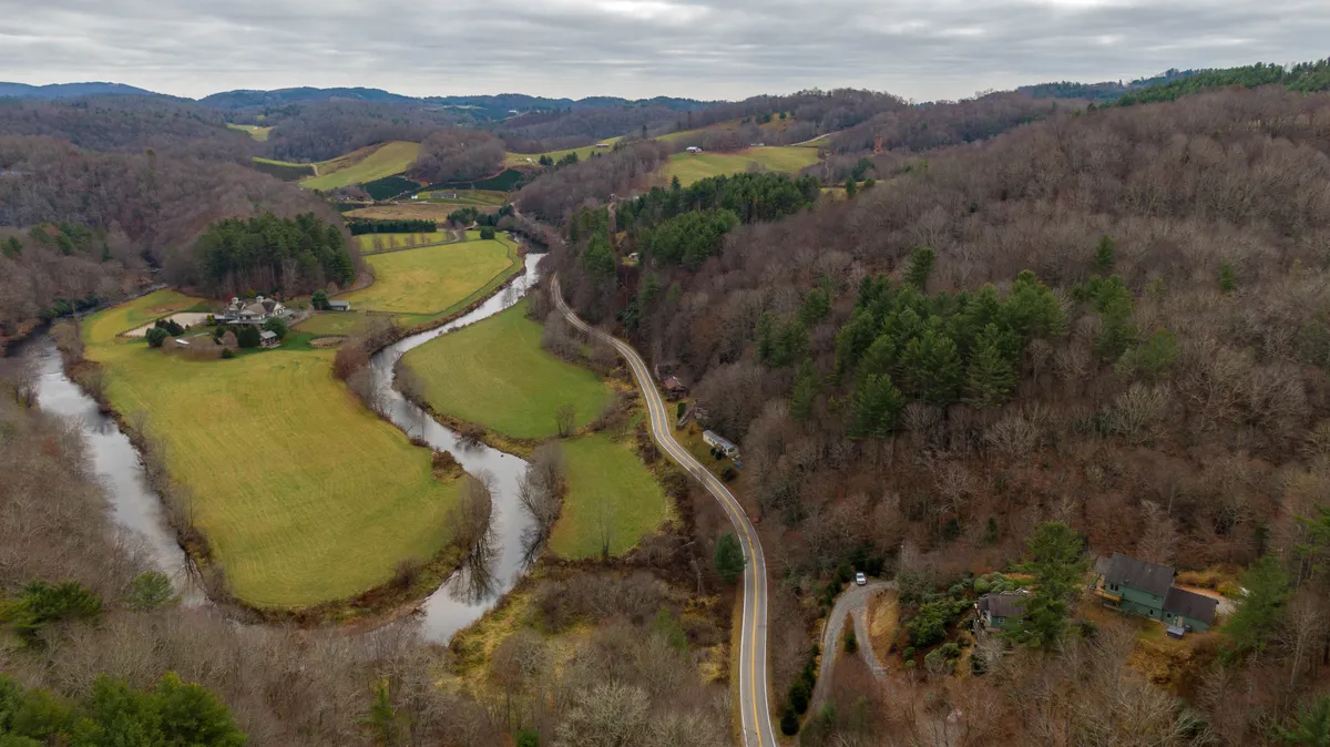 A sweeping view of a river gently winding through verdant fields, framed by dense forested hills and dotted with scattered homes. The scene radiates tranquility and adventure, suggesting endless possibilities for fishing, hiking, and exploring the Appalachian landscape.
