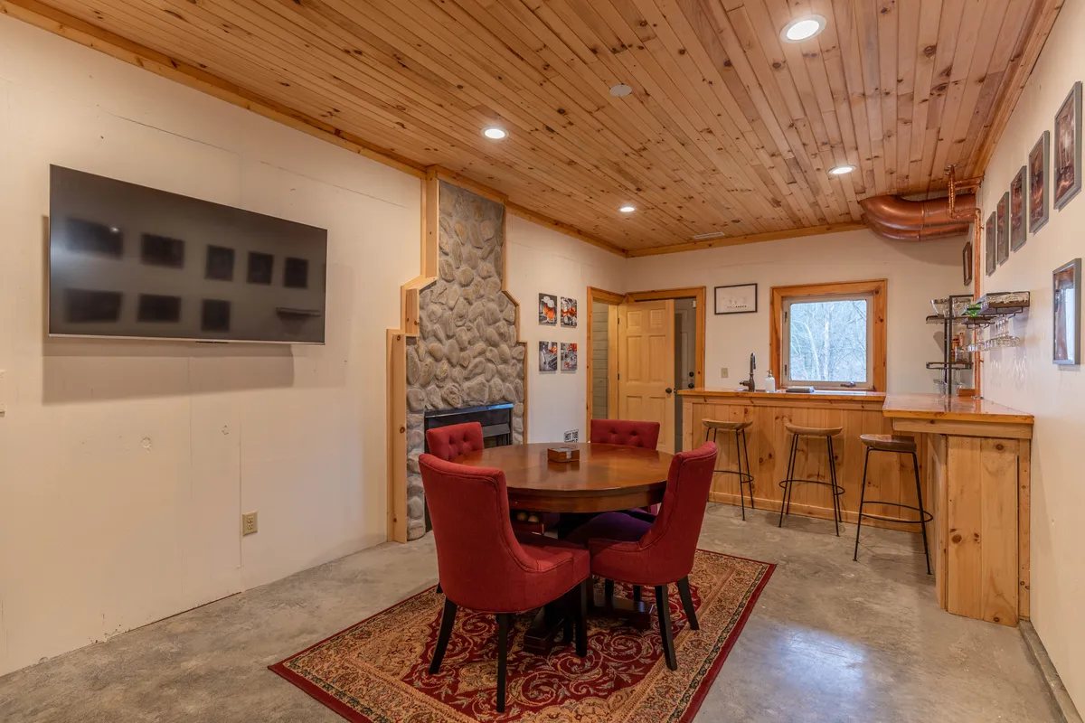 A well-appointed entertainment space showcasing a round wooden table surrounded by red chairs, a wall-mounted TV, a stone fireplace, and a bar area with stools. The setup suggests a place for both spirited games and relaxing social moments.