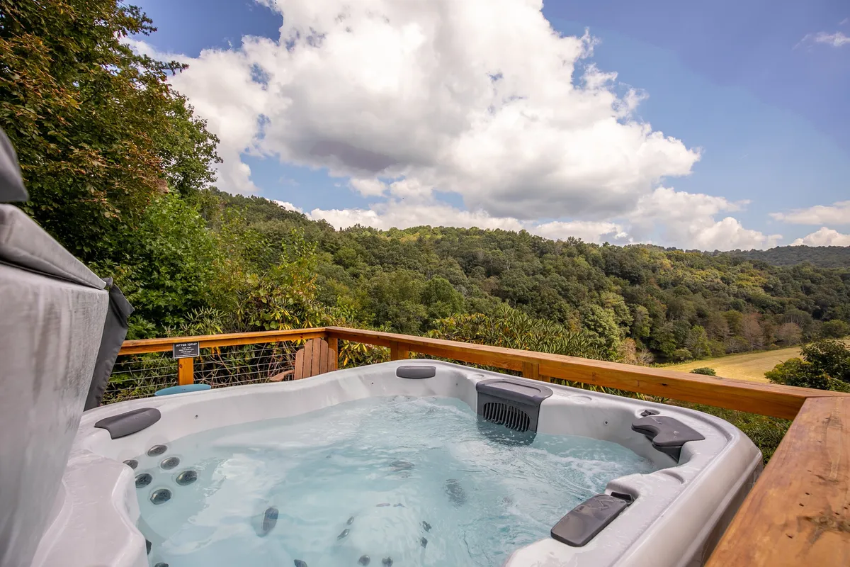 A hot tub sits on a wooden deck overlooking a scenic landscape, bordered by a wooden railing. The surrounding view is lush and open, inviting guests to unwind amid nature.