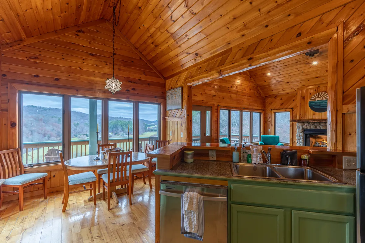 This image features a spacious dining area with a large table set for six, adjacent to a modern kitchen island, and walls of windows that allow in abundant natural light and frame scenic views. Hand-crafted woodwork and hardwood floors are visible, with a seamless flow between the kitchen and dining spaces, emphasizing the open-concept design.