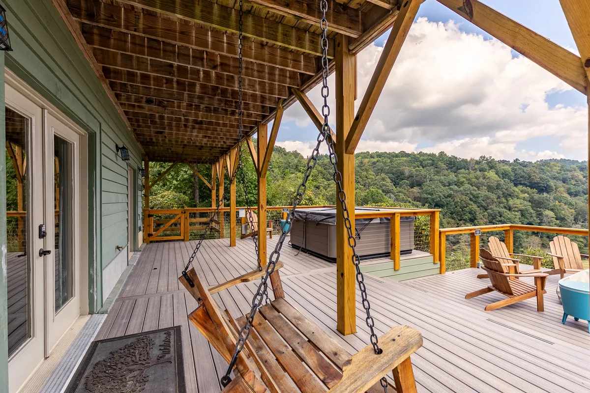 This image captures a spacious wooden deck featuring a cozy swinging bench, an inviting hot tub, Adirondack chairs, and lush green views. The deck is designed for both relaxation and socializing, with the swing and hot tub positioned to take in the surrounding scenery.