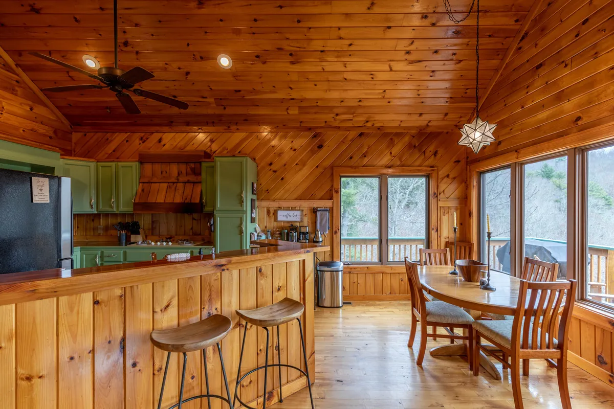 The shot centers on a modern kitchen island and dining table, surrounded by large windows that overlook breathtaking scenery. The hardwood floors and custom cabinetry reflect the artisan craftsmanship, while the setup invites both cooking and conversation.