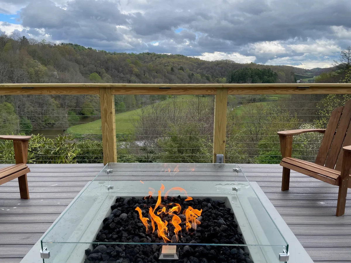 A cozy outdoor fire pit area on a wooden deck, with Adirondack chairs arranged for socializing against the backdrop of a stunning mountain view. The space is clearly designed for evening gatherings, roasting marshmallows, and stargazing.