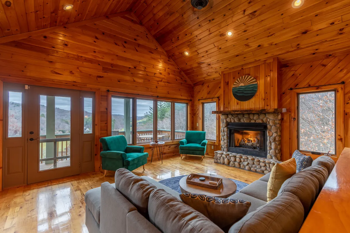A cozy living room with blue chairs arranged near a substantial stone fireplace, surrounded by wooden walls and wide windows offering sunset views. The space is designed for comfort, with thoughtful decor and an atmosphere that encourages relaxation and group conversations.