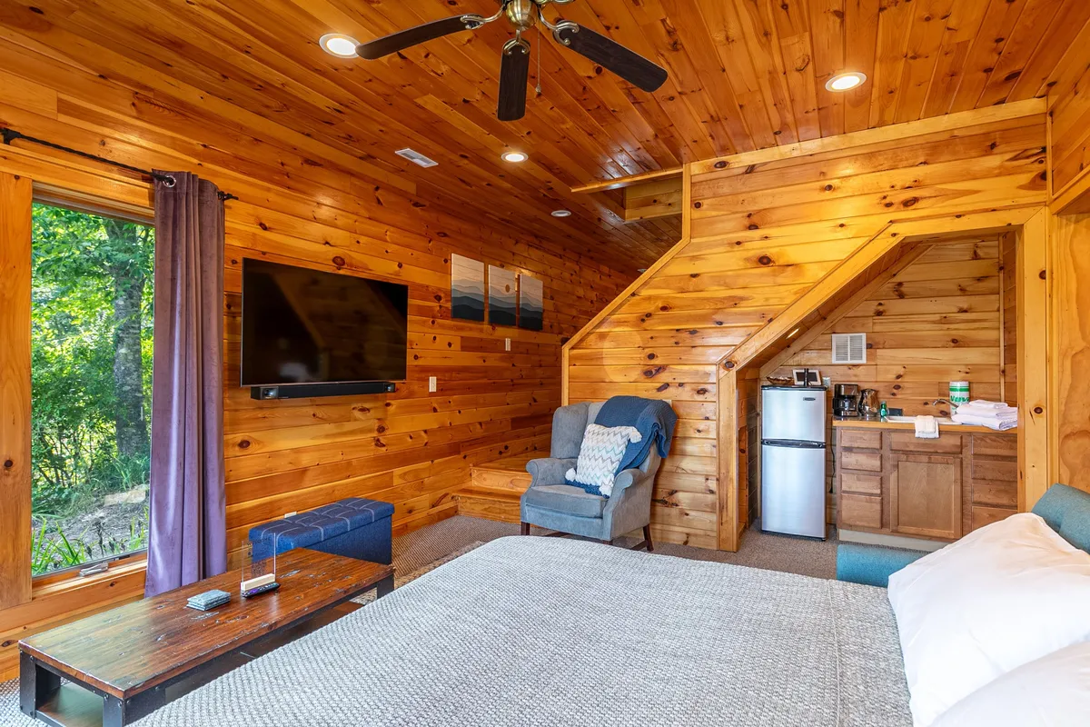A warm and inviting bedroom interior featuring a king-sized bed, wooden walls, armchair seating, and a television. There's a window that suggests views of the outdoors, contributing to a sense of privacy and coziness.