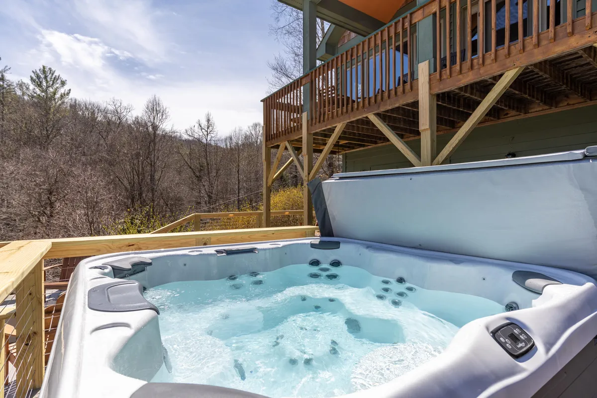 A wooden deck nestled in lush greenery features a bubbling hot tub surrounded by nature. The deck offers ample space for relaxing, with an inviting view of the surrounding forest, perfect for unwinding under the stars or enjoying a soak in the fresh mountain air.