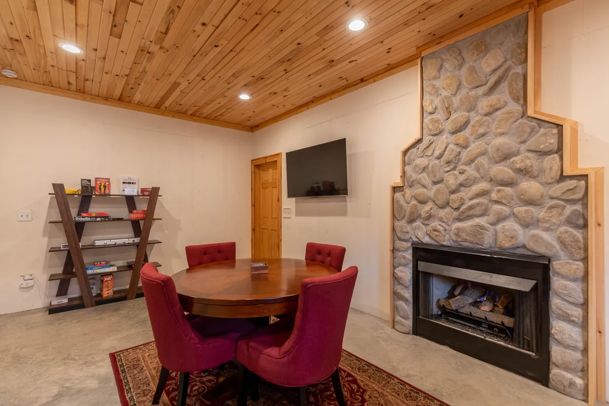 A warmly-lit game and lounge area featuring a round wooden table with red upholstered chairs set near a stone fireplace. The room includes shelves stocked with books and board games, a flat-screen TV for media, and cozy decor that invites group gatherings and evening fun.